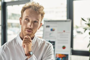 Kızıl saçlı odaklanmış bir işadamı, çağdaş ofis alanındaki bir projede sonraki adımlarını düşünüyor..