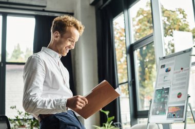 Beyaz gömlekli neşeli bir kızıl doğal ışık dolu çağdaş bir ofiste proje belgelerini inceliyor..
