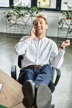 A red-haired businessman in a white shirt happily discusses a project on his phone while relaxing in an office. clipart