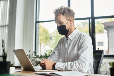 Beyaz tişörtlü kızıl saçlı bir işadamı, bir proje üzerinde çalışıyor. Çağdaş bir ofis alanında, maske takıyor..