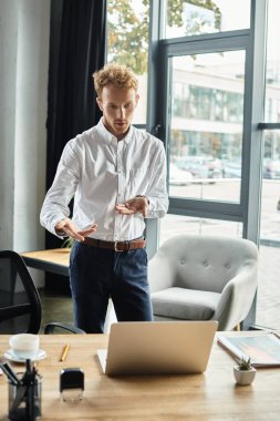 A focused redhead businessman in a white shirt discusses project ideas at his desk in a contemporary office. clipart