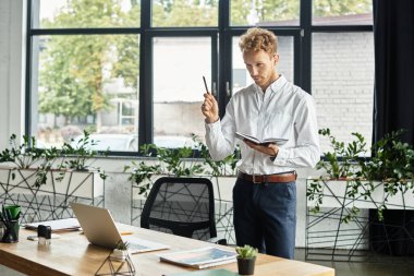 A focused businessman with red hair stands with a notebook, passionately brainstorming ideas at his desk. clipart