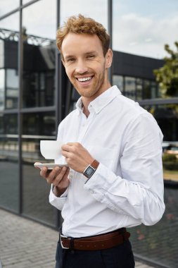 Beyaz gömlekli neşeli bir kızıl kahve fincanı tutuyor. Ofisteki bir iş projesini düşünüyor..