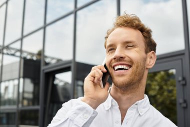 A cheerful businessman with red hair discusses a project on his phone while in a sleek office. clipart