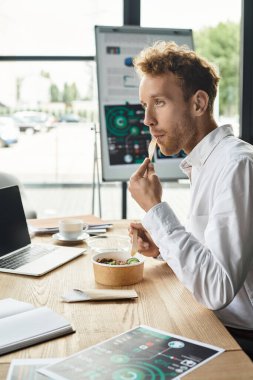 Kızıl saçlı odaklanmış bir işadamı şık bir ofis alanında işine konsantre olurken yemeğin tadını çıkarıyor..