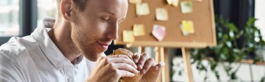 A focused businessman with red hair relishes a burger in his contemporary office while working on a project. clipart