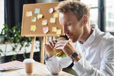 A redhead businessman pauses to savor a burger during a creative work session in his stylish office. clipart