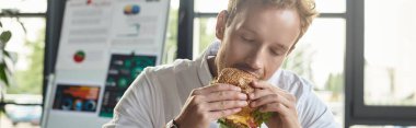 Profesyonel bir kızıl burgerini ısırıyor. Parlak bir ofis ortamından memnun ve nişanlı..