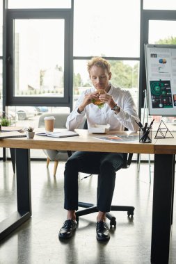 A redhead businessman in a white shirt focuses on lunch while involved in a work project at a sleek office. clipart