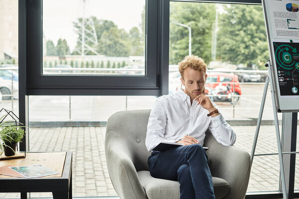 A redheaded man in a white shirt reflects on his project while seated in a chic office chair.