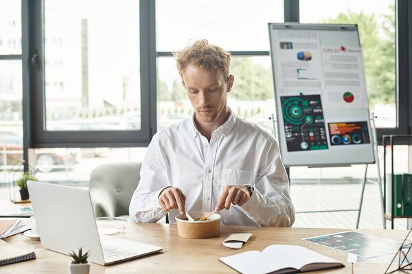 A determined redhead businessman in a white shirt works on a project while enjoying lunch at his desk.