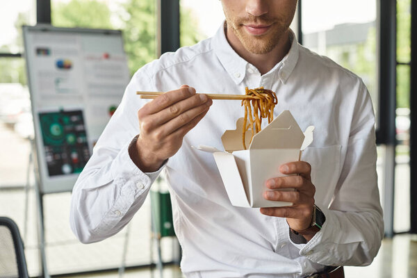 A focused businessman in a white shirt eats noodles with chopsticks during his productive workday.