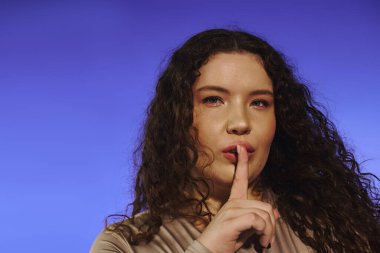 Kendine güvenen, kıvırcık saçlı, kendine güvenen bir kadın. Sessizlik için şakacı hareketler yapar..