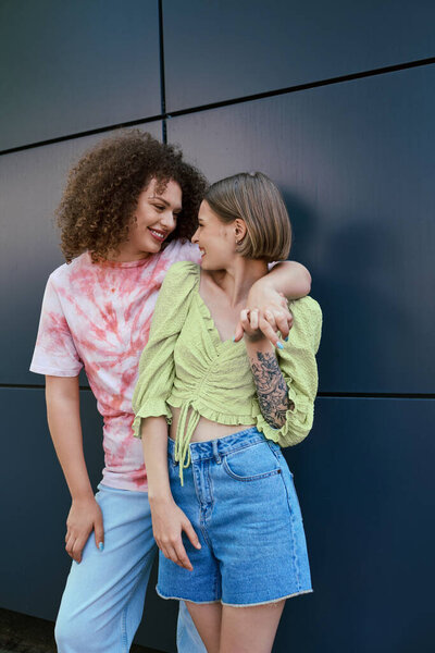 Две женщины разделяют смех и любовь, стоя вместе на современном фоне, излучая радость.