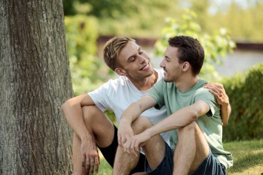 Two smiling men sit close together in a lush park, sharing laughter and affection. clipart