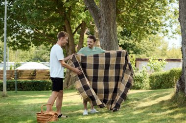 Two joyful men unfold a blanket together in a sunlit park, preparing for a delightful picnic. clipart