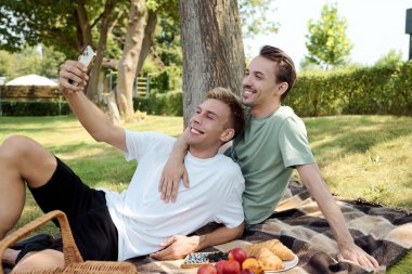 Two men enjoy a delightful picnic, smiling while taking a selfie together in a serene park. clipart