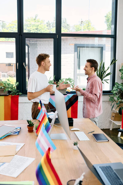 Двое улыбающихся людей наслаждаются сердечным разговором, украшенным флагами гордости в солнечном офисе.