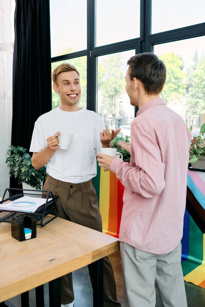 Двое веселых мужчин беседуют за чашечкой кофе в оживленной офисной атмосфере.