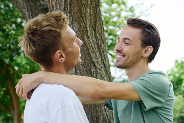 Two affectionate men share a loving embrace under a tree, smiles lighting up their faces.