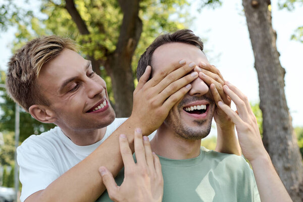 Два улыбающихся мужчины разделяют игривый момент на открытом воздухе, окруженный зеленью и солнцем.