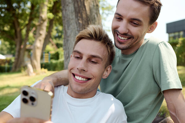 Веселая гей-пара улыбается на телефон, разделяя восхитительный момент в парке.