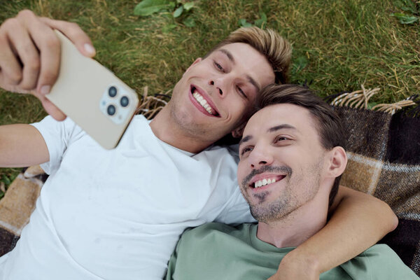 A handsome couple enjoys a sunny day, smiling while taking a selfie together on a blanket.