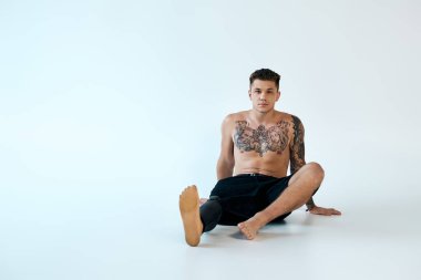 A confident young man with tattoos sits on the floor, showcasing his prosthetic leg in a well-lit studio. clipart