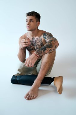 A young man with tattoos poses in a studio, highlighting his prosthetic leg and muscled form while deep in thought. clipart