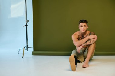 Çarpıcı dövmeleri olan genç bir adam stüdyo zemininde oturarak protez bacağını ön plana çıkarıyor..