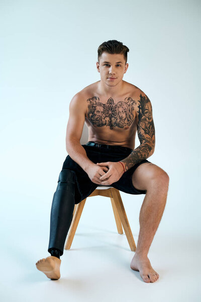 Татуированный молодой человек показывает свою протезную ногу, сидя на стуле в гладкой студийной обстановке.