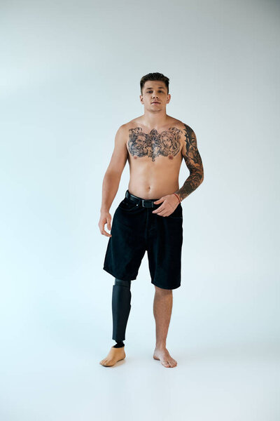 Молодой человек с яркими татуировками уверенно стоит, показывая свою протезную ногу в минималистской студии.