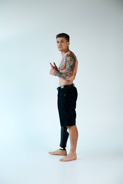 Татуированный молодой человек гордо стоит в студии, демонстрируя свою протезную ногу и яркие татуировки..