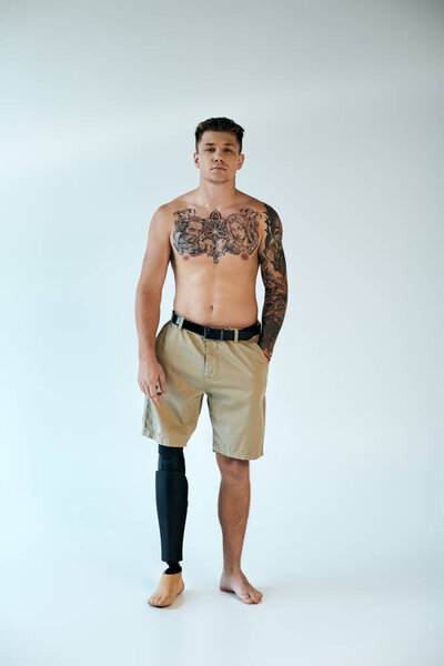 Татуированный молодой человек с протезной ногой гордо стоит в студии, излучая уверенность и силу.