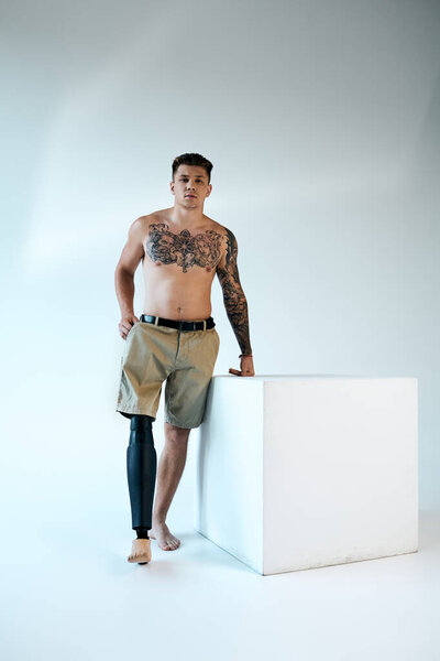 Уверенный в себе молодой человек с татуировками гордо стоит в студии, демонстрируя свою протезную ногу и силу.