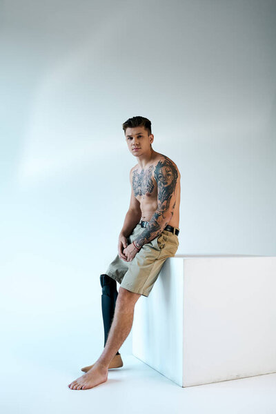 Уверенный в себе молодой человек с татуировками сидит в студии, демонстрируя свою протезную ногу и художественное выражение.