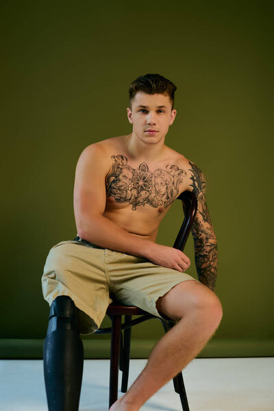 Татуированный молодой человек сидит на стуле, раскрывая свою протезную ногу и демонстрируя свое тело в студии..
