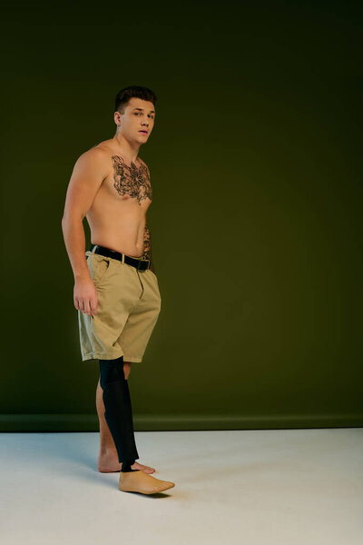 Татуированный молодой человек гордо стоит в студии, подчеркивая свою уникальную протезную ногу и художественный дух..