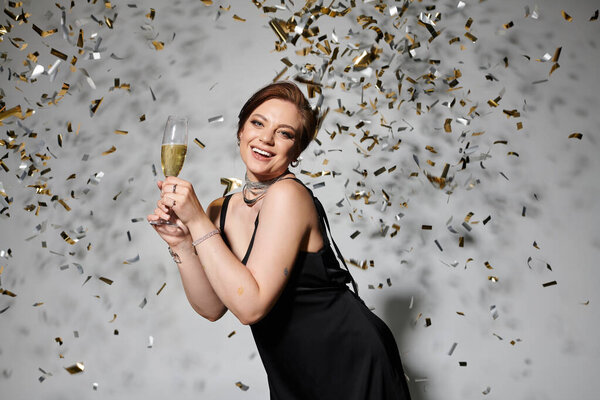 A stylish woman raises a champagne glass and smiles joyfully as golden confetti falls around her.