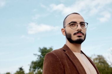 Şık bir sonbahar kıyafeti giymiş kendine güvenen bir adam canlı bir sonbahar yeşilliği arasında duruyor, doğanın güzelliğini kucaklıyor..