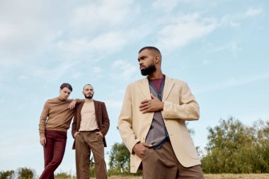 Three fashionable men confidently pose amidst golden autumn scenery, highlighting seasonal trends. clipart