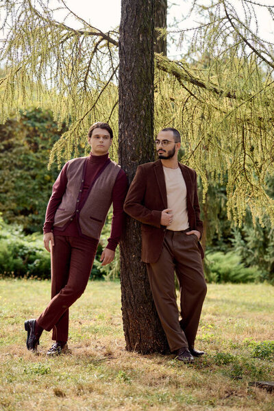 Двое красивых мужчин, одетых в модный осенний наряд, уверенно стоят в красивом поле, окруженном природой.