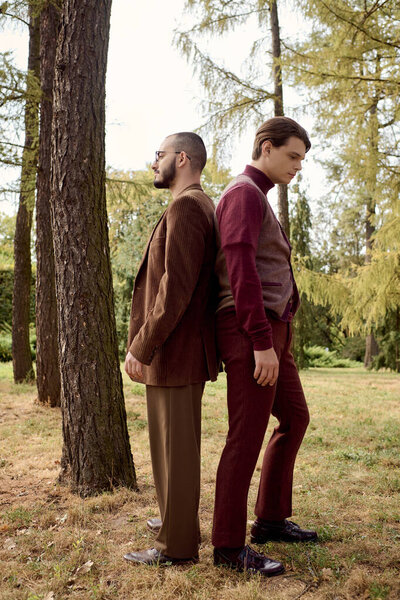 Two handsome men dressed in autumn fashion stand back to back in a tranquil natural setting.