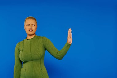 Genç bir kadın kendine güveni tam yeşil bir elbise içinde mavi bir arka plana karşı poz veriyor..