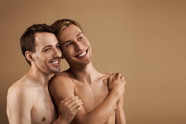 Two young men embrace closely, smiling warmly in a cozy, neutral toned environment.