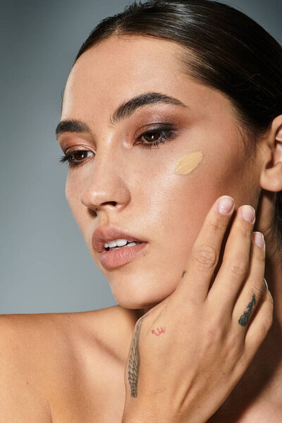 Потрясающая женщина касается ее щеки, когда она применяет фундамент, демонстрируя свою красоту в мягком свете.