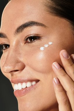 A woman with radiant skin gracefully applies cream to her cheek while smiling. clipart