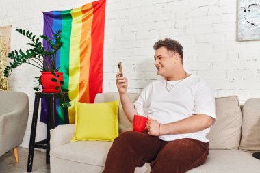 Gülümseyen bir birey kırmızı bir bardak tutuyor ve telefonlarına bakıyor, renkli dekorlarla çevrili..