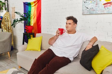 A unique individual enjoys a drink in a cozy, colorful environment adorned with a rainbow flag clipart