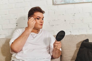 A queer person carefully enhances their appearance using makeup while sitting on a comfortable sofa. clipart